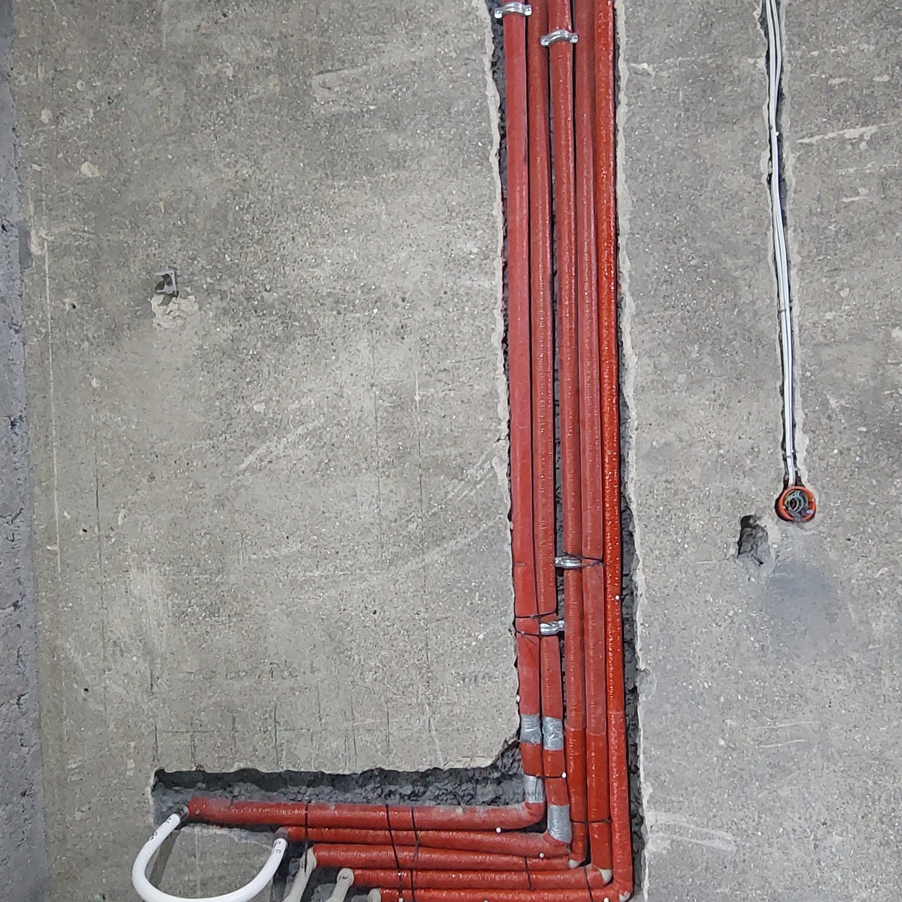 Czerwone rury ogrzewania podłogowego, mocowane do surowej ściany z betonu, z widoczną instalacją elektryczną. Etap prac instalacyjnych przed wylewką.