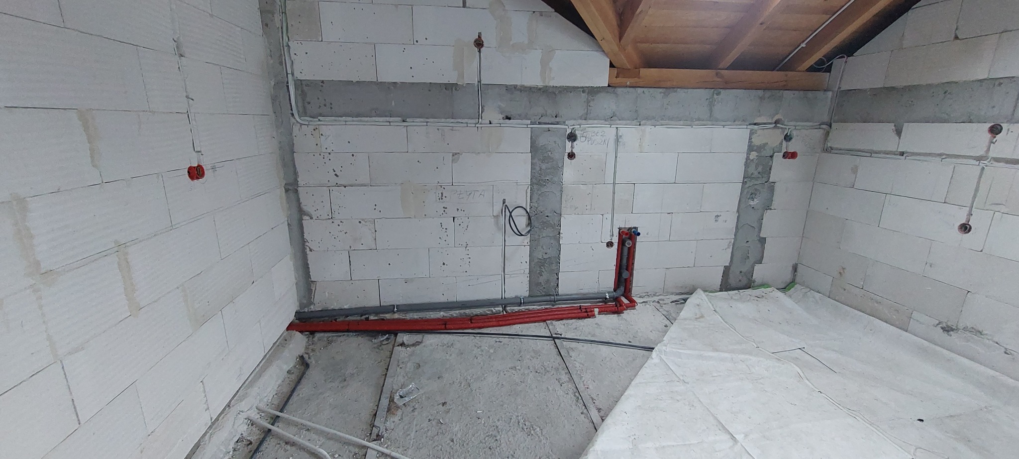 Wnętrze w trakcie prac instalacyjnych: widoczne przewody elektryczne, rury grzewcze i wodne na tle surowych ścian z betonu komórkowego i drewnianej konstrukcji dachu.
