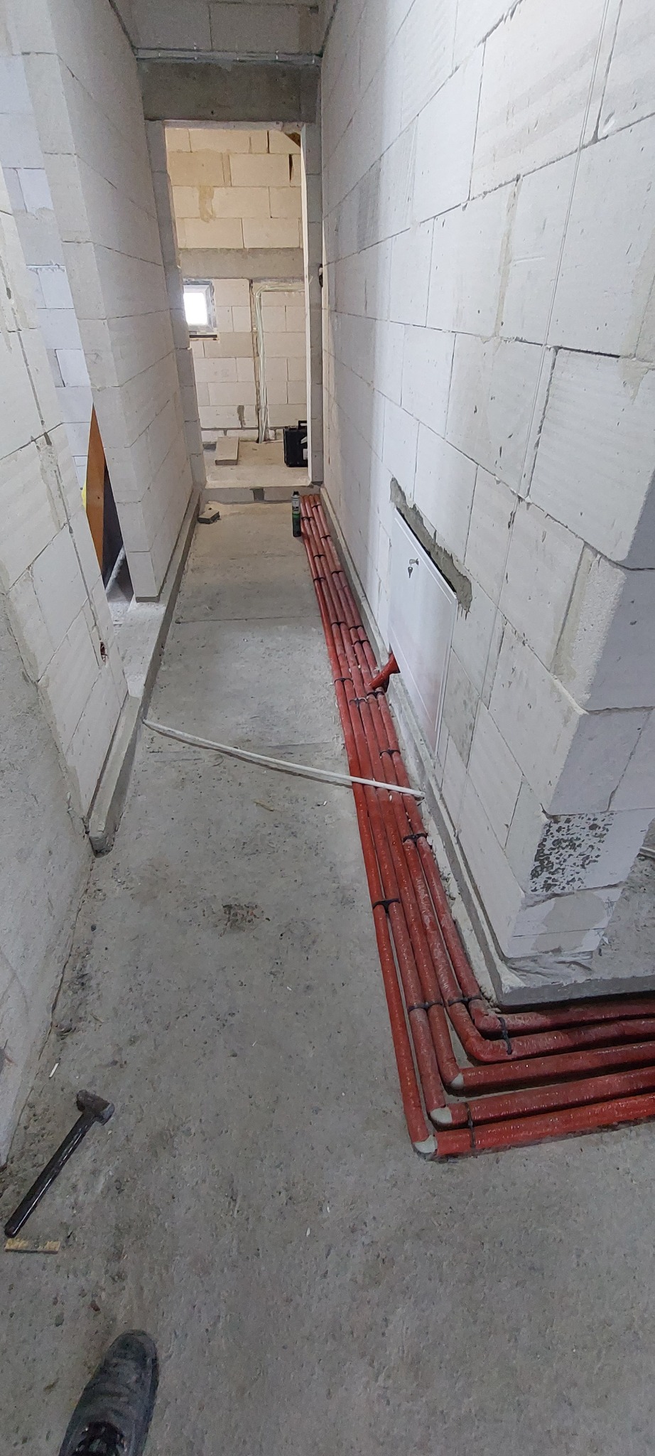 Czerwone rury ułożone wzdłuż ściany z białych bloczków w surowym wnętrzu budynku. Widoczny fragment podłogi i narzędzie (młotek).