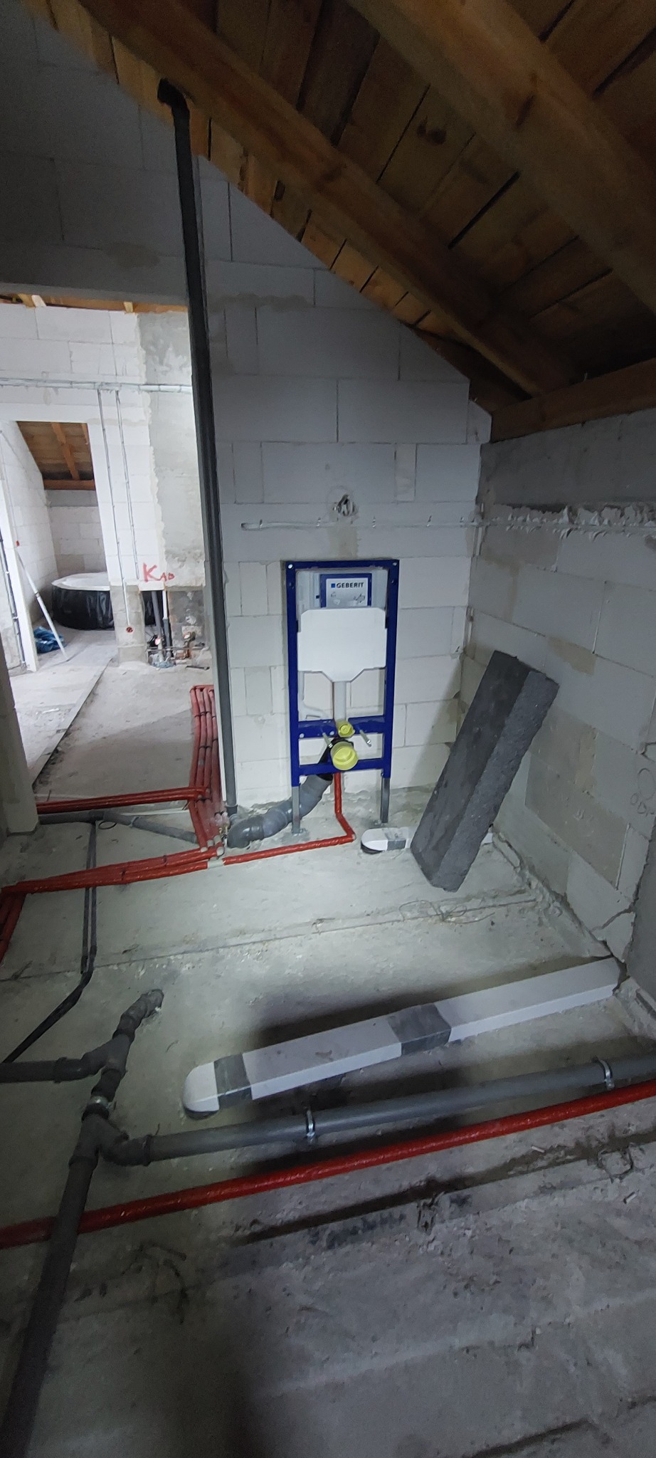 Instalacja podtynkowa Geberit w surowym wnętrzu poddasza. Widoczne rury instalacyjne, elementy montażowe i fragment drewnianej konstrukcji dachu.