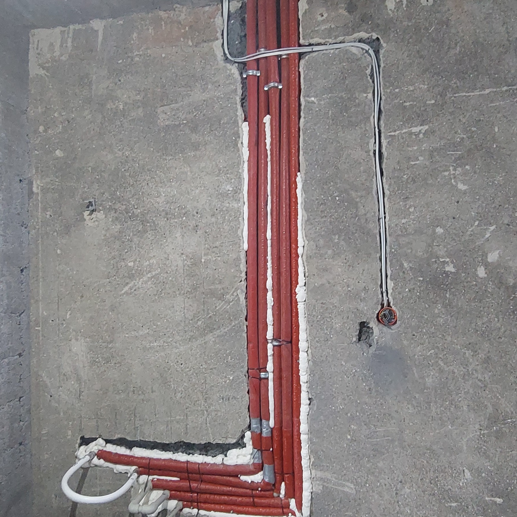 Czerwone rury w bruzdach ściany, zabezpieczone pianką montażową i uchwytami. Widoczne przewody elektryczne i surowy beton. Instalacja w trakcie prac.