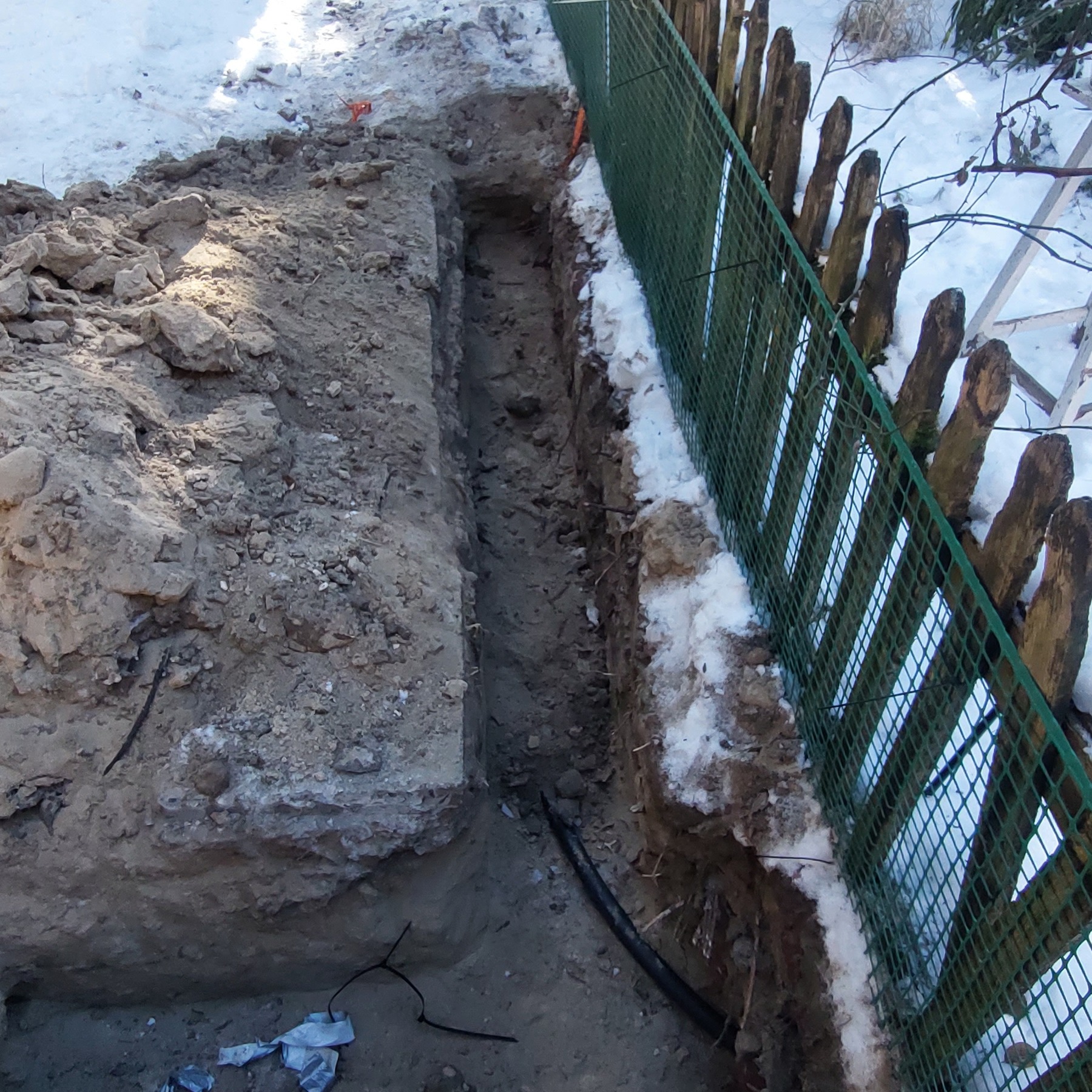 Wąski wykop w ziemi, częściowo pokrytej śniegiem, biegnący wzdłuż ogrodzenia z siatki. Widoczne kable w wykopie. Ziemia gliniasta, grudkowata. Słoneczny dzień.