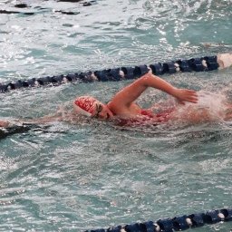 LINALO Karolina Pawera - Kobieta w czerwonym stroju i czepku z płomieniami płynie kraulem w basenie, tworząc fale i pianę. Widoczna lina oddzielająca tory.