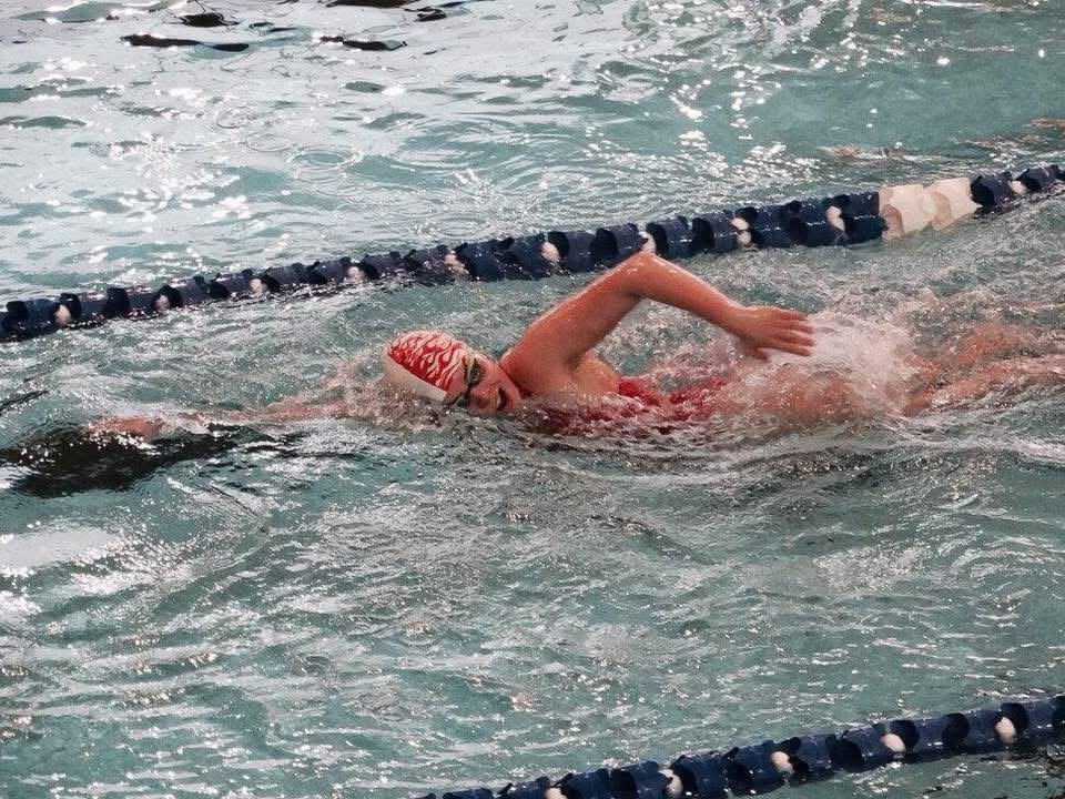 Kobieta w czerwonym stroju i czepku z płomieniami płynie kraulem w basenie, tworząc fale i pianę. Widoczna lina oddzielająca tory.