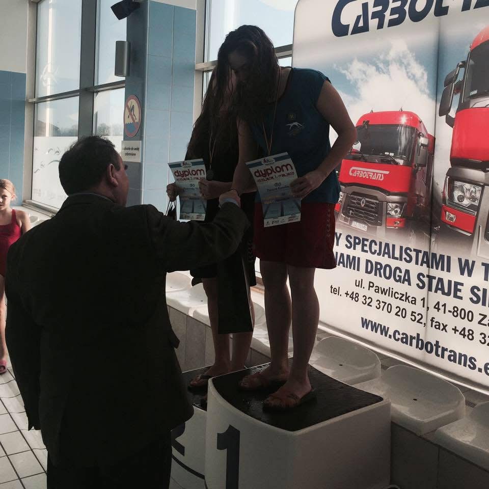 Dekoracja zawodniczek na podium w hali basenowej. Wręczenie dyplomów za zawody pływackie. W tle baner reklamowy sponsora zawodów.