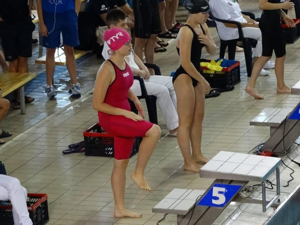 Pływaczka w czerwonym stroju robi rozgrzewkę na bloku startowym z numerem 5, w tle inni zawodnicy i basen. Przygotowania do zawodów pływackich.