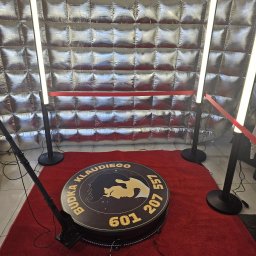 Budkaklaudiego360 - Okrągła fotobudka z logiem na czerwonym dywanie, oświetlona lampami LED, na tle srebrnej, pikowanej ścianki. Widoczne okablowanie i elementy montażowe.