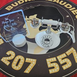 Budkaklaudiego360 - Retro biały telefon i podświetlona instrukcja 'Audio Księga Gości' na okrągłym podeście z napisem 'Budka Klaudiego 360' i numerem telefonu.