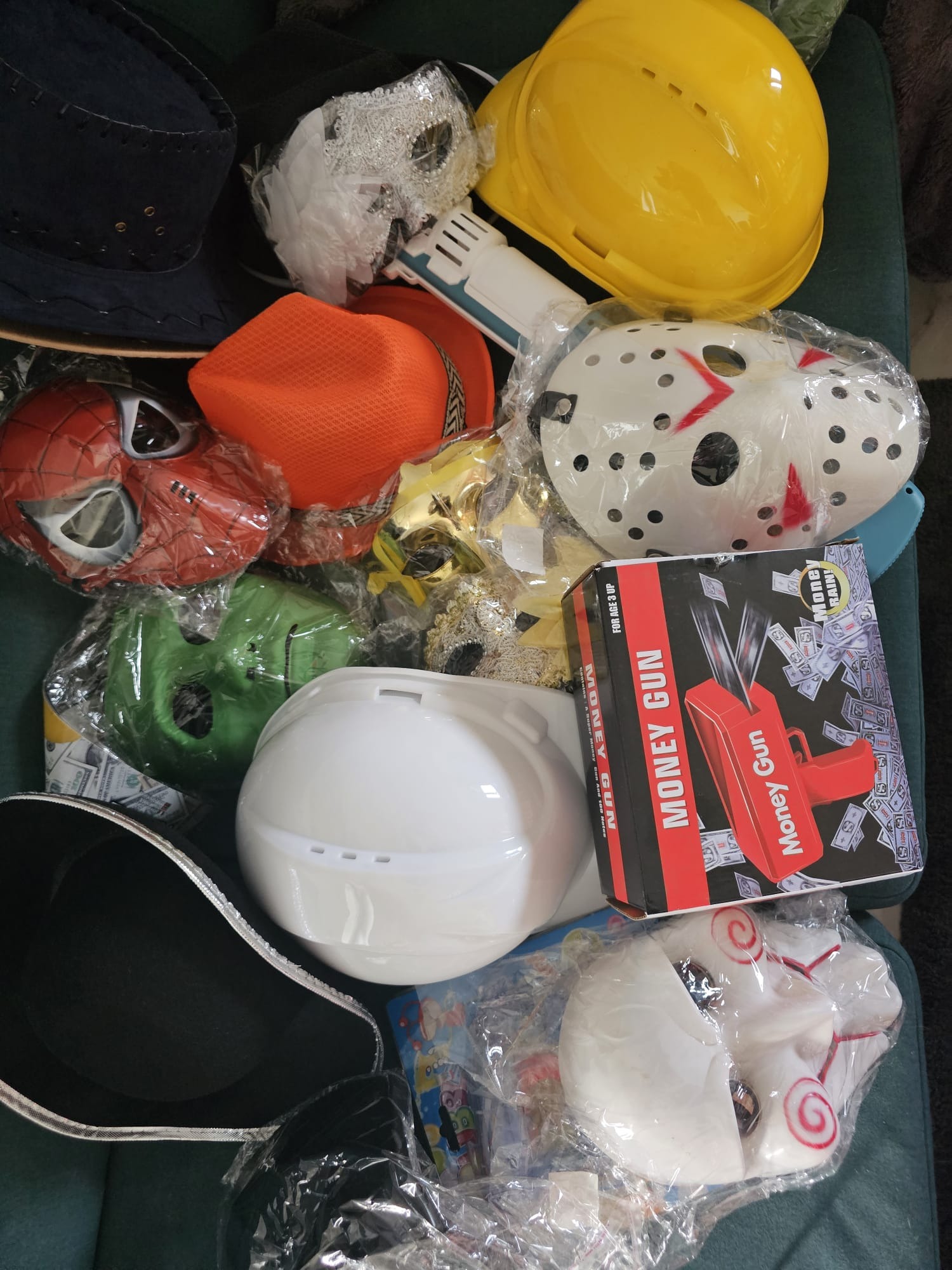 Kolekcja masek i rekwizytów imprezowych: maski Spider-Mana, Jasona, zielona maska, kapelusze, kask i pudełko 'Money Gun' na zielonej powierzchni.