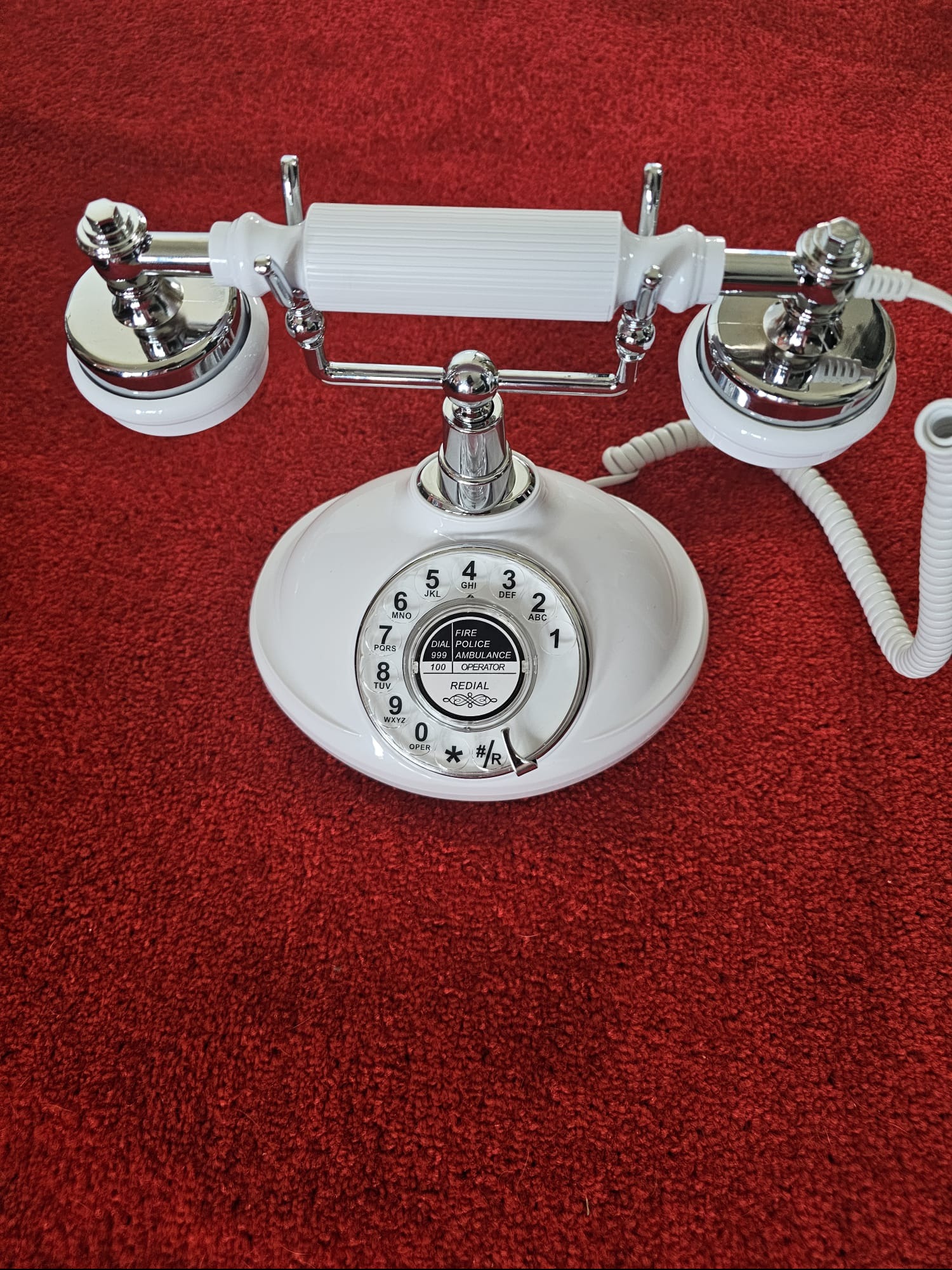 Biały, retro telefon stacjonarny z chromowanymi elementami na czerwonym tle. Centralny widok, lekko z góry. Widoczne numery i napis 'DIAL POLICE'.