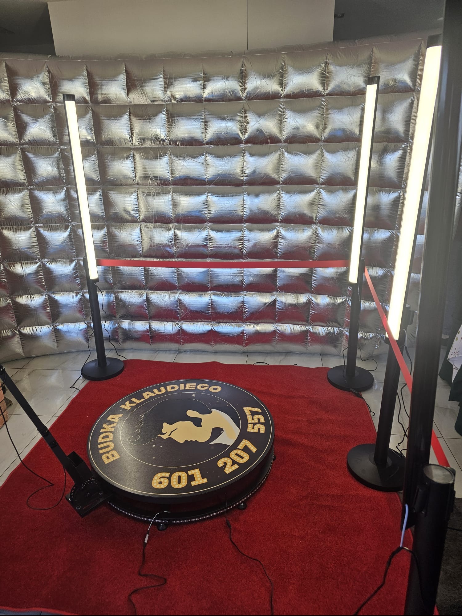 Oświetlona fotobudka 360 na czerwonym dywanie, z srebrnym, pikowanym tłem i numerem telefonu. Budka Klaudiego, gotowa do użycia na imprezie.