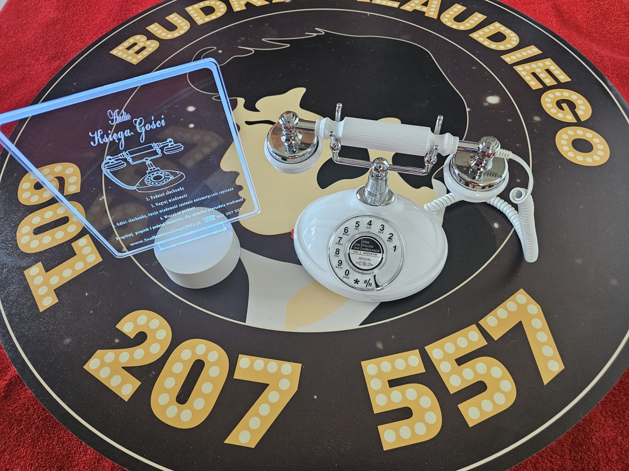 Retro biały telefon i podświetlona instrukcja 'Audio Księga Gości' na okrągłym podeście z napisem 'Budka Klaudiego 360' i numerem telefonu.