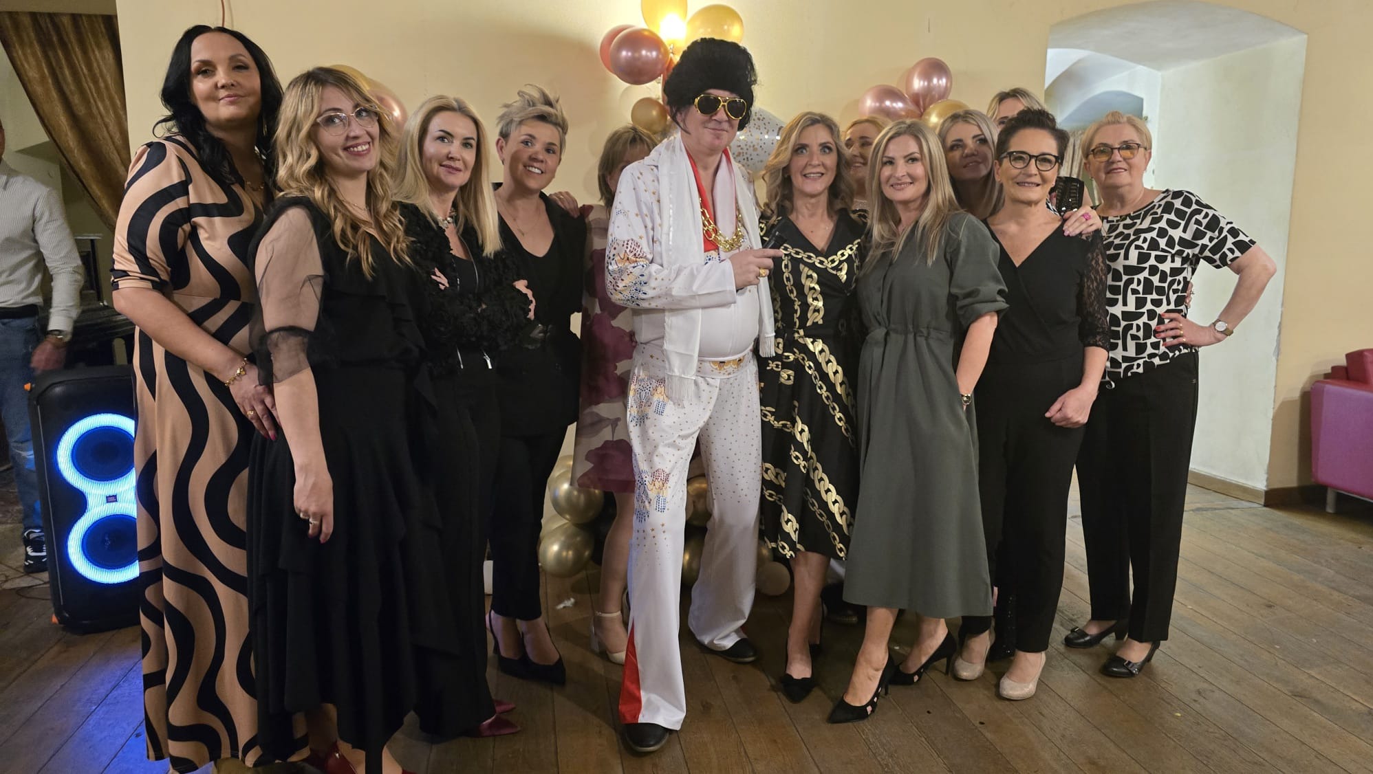 Elvis Presley tribute artist, w otoczeniu kobiet, pozuje na tle balonów podczas imprezy. Widoczny głośnik JBL.