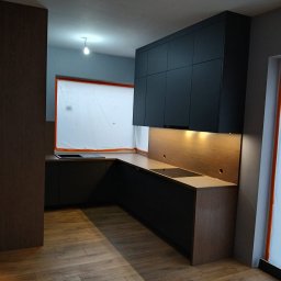 STOLMAR - Nowoczesna kuchnia z czarnymi szafkami górnymi i drewnianymi blatami. Widoczne oświetlenie podszafkowe i drewniana podłoga. Okno zabezpieczone folią malarską.