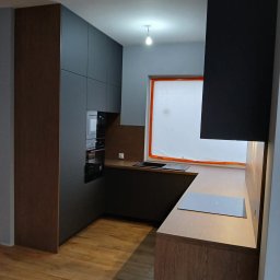 STOLMAR - Nowoczesna kuchnia w odcieniach szarości i drewna z zabudowanymi sprzętami AGD, oświetleniem punktowym i drewnianą podłogą. Okno zabezpieczone folią malarską.