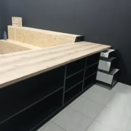 Nowoczesna zabudowa meblowa z blatem imitującym drewno, czarne półki i wysuwane białe szuflady. Ujęcie z perspektywy podłogi, widoczna butelka wody.