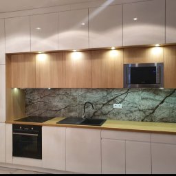 STOLMAR - Nowoczesna kuchnia z białymi frontami i drewnianymi szafkami górnymi, podświetlona. Blat drewniany, na ścianie dekoracyjny panel imitujący marmur.