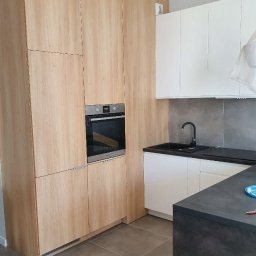 STOLMAR - Jasna, drewniana zabudowa kuchenna z piekarnikiem w zabudowie, kontrastująca z białymi szafkami i ciemnym blatem. Nowoczesne wykończenie, szare płytki na ścianie.