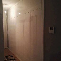 STOLMAR - Biała szafa wnękowa z połyskiem, zabudowa od podłogi do sufitu w przedpokoju. Na podłodze dywanik, obok szarej ściany wisi regulator temperatury.