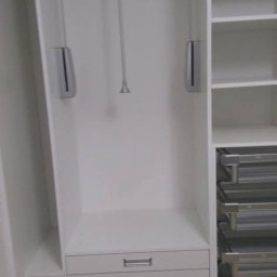 STOLMAR - Wnętrze białej szafy wnękowej z wysuwanymi półkami i drążkiem na ubrania. Widoczne trzy szuflady z metalowymi uchwytami i półki z metalowymi koszami.