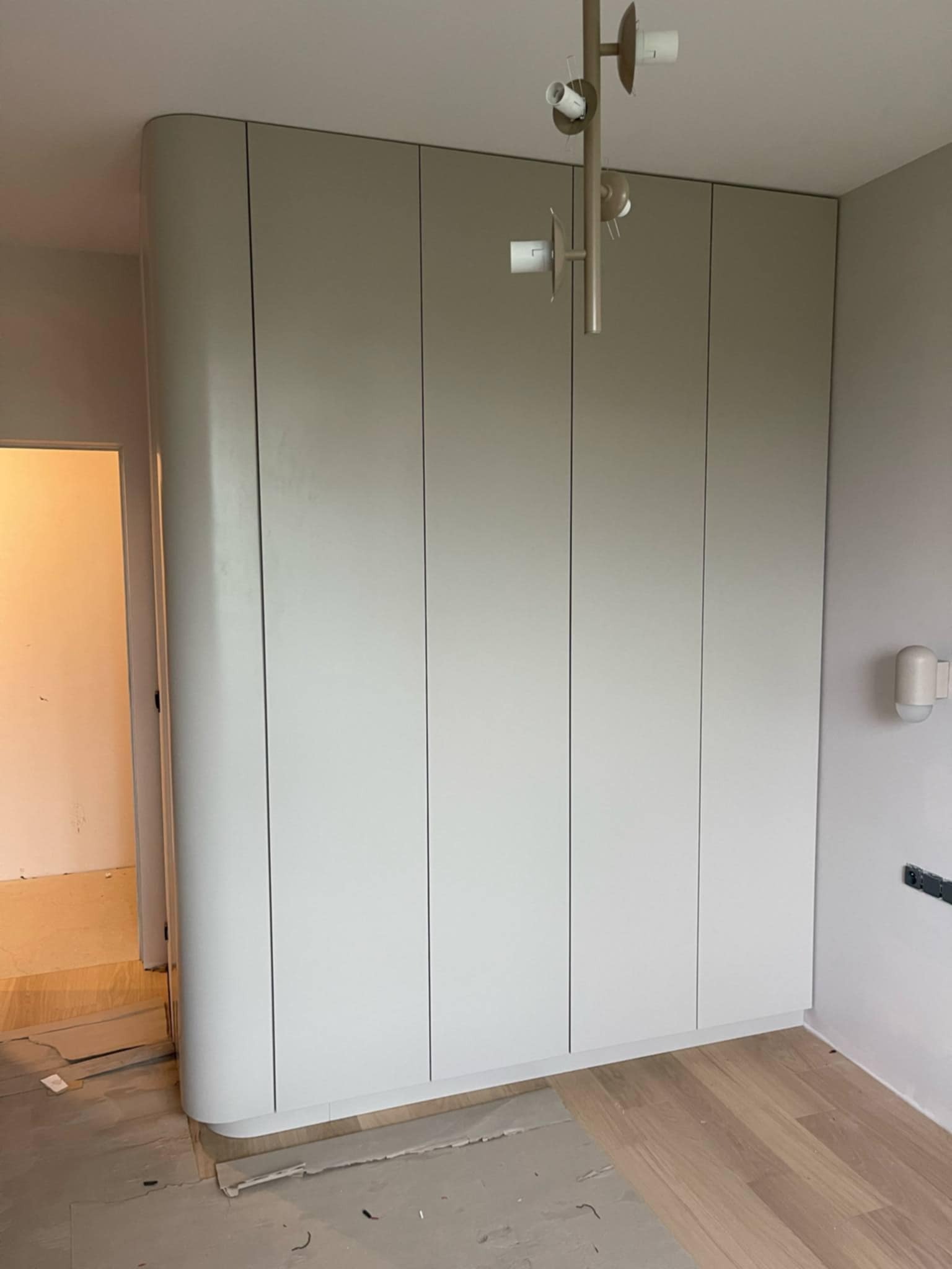 Jasna szafa wnękowa z zaokrąglonym bokiem, w trakcie montażu, widoczne niedokończone podłogi i lampa sufitowa. Minimalistyczny design.
