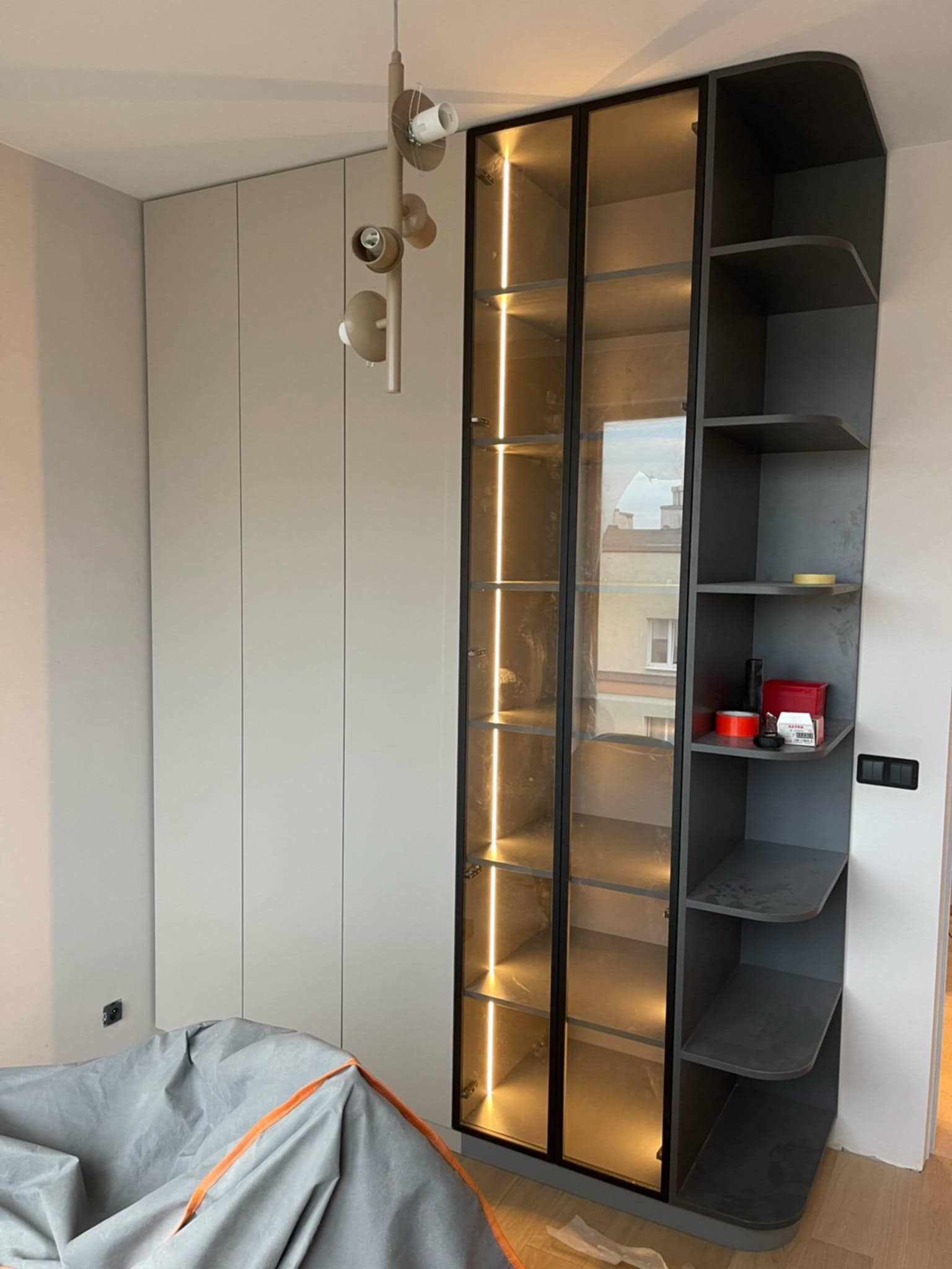 Nowoczesna szafa wnękowa z szklanymi drzwiami i podświetleniem LED, z półkami w odcieniach szarości, widoczna w trakcie montażu, przykryta folią ochronną.