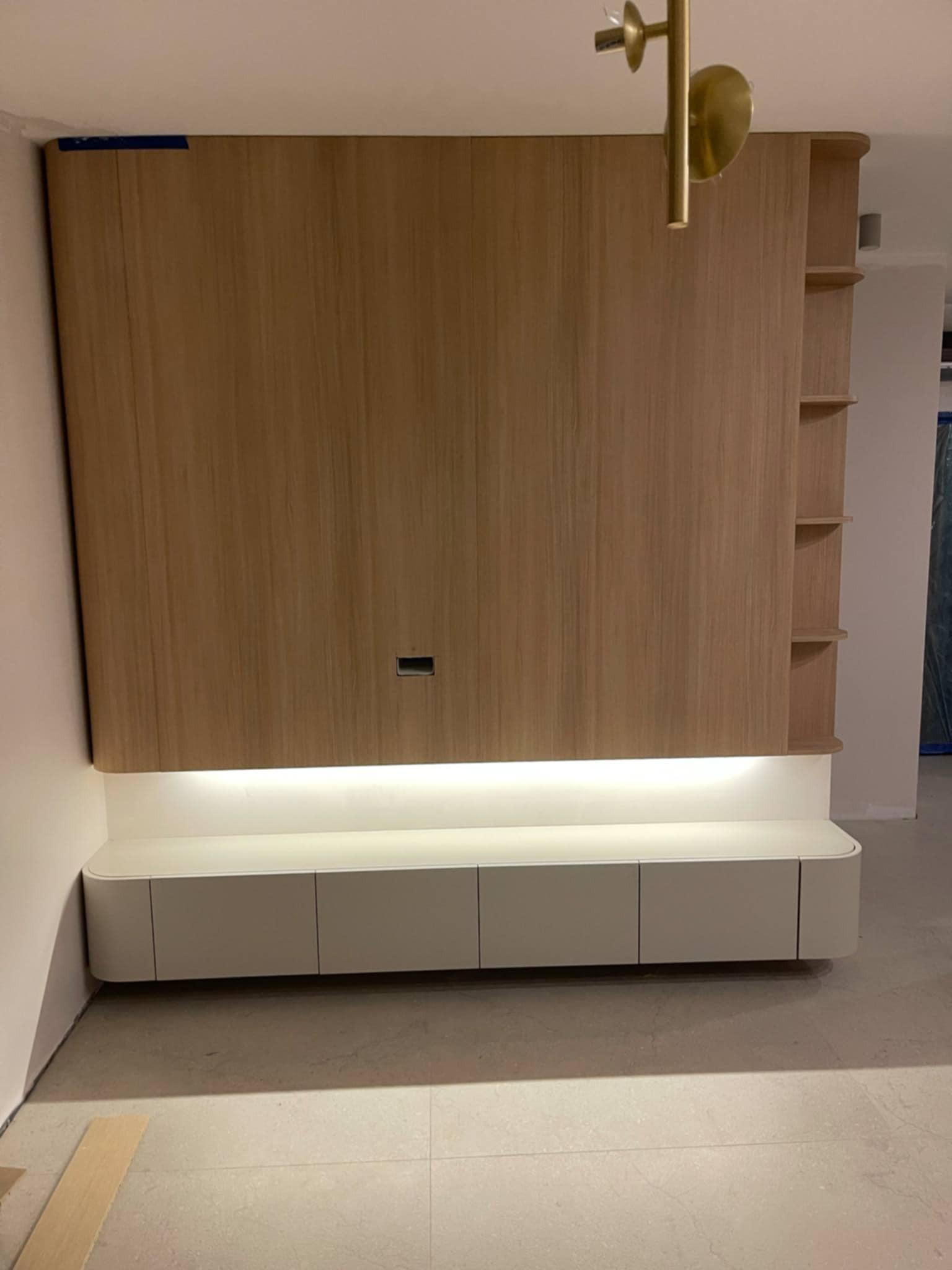 Drewniana zabudowa ściany z szafkami i półkami, podświetlona od dołu. Widoczne okleiny na krawędziach i lampa sufitowa. Minimalistyczny design.