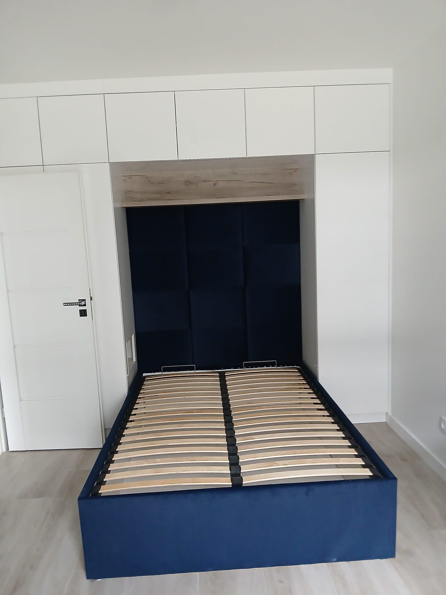 Niebieskie łóżko tapicerowane ze stelażem, wbudowane w białą zabudowę meblową z szafkami. Wezgłowie z paneli tapicerowanych. Minimalistyczne wnętrze.