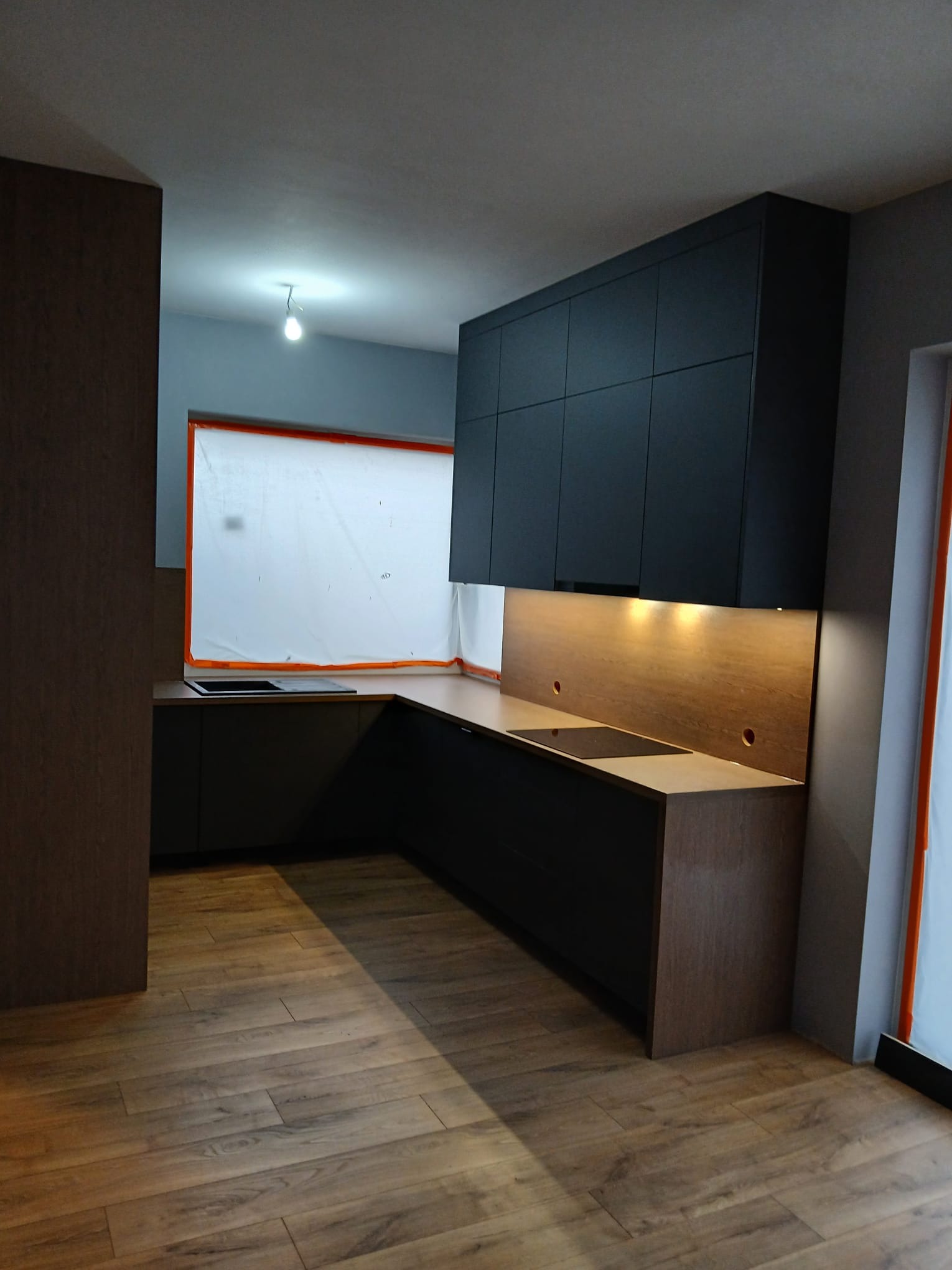 Nowoczesna kuchnia z czarnymi szafkami górnymi i drewnianymi blatami. Widoczne oświetlenie podszafkowe i drewniana podłoga. Okno zabezpieczone folią malarską.