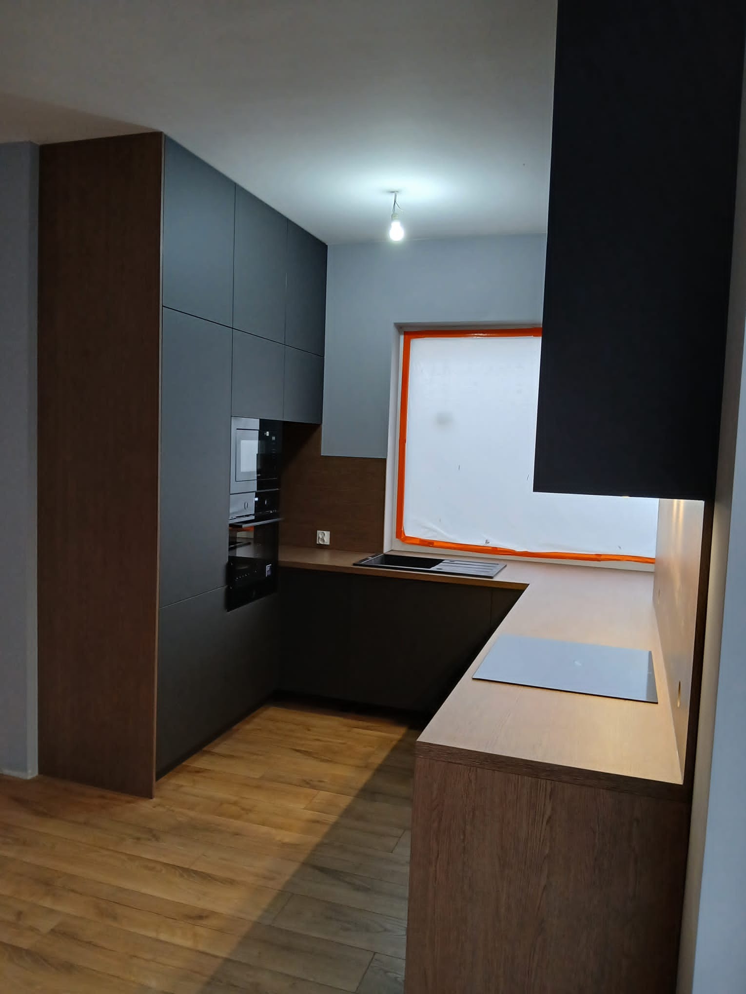 Nowoczesna kuchnia w odcieniach szarości i drewna z zabudowanymi sprzętami AGD, oświetleniem punktowym i drewnianą podłogą. Okno zabezpieczone folią malarską.