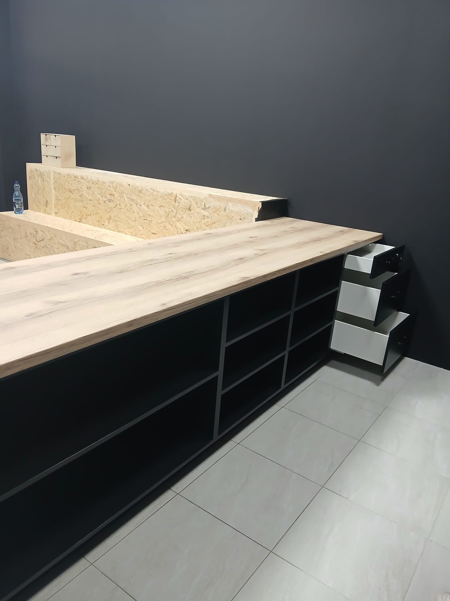 Nowoczesna zabudowa meblowa z blatem imitującym drewno, czarne półki i wysuwane białe szuflady. Ujęcie z perspektywy podłogi, widoczna butelka wody.