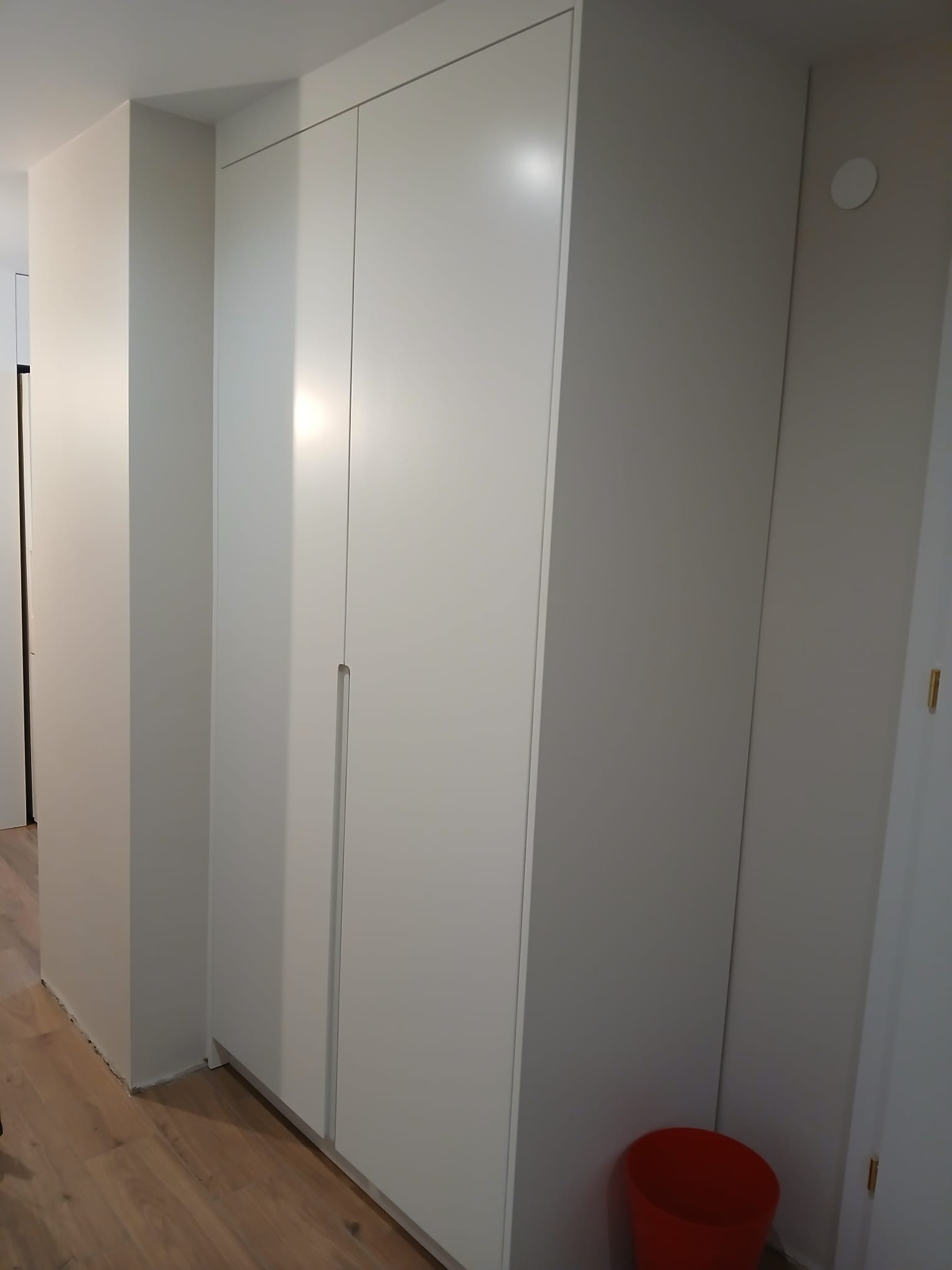 Wnętrze z zabudowaną, białą szafą wnękową z subtelnymi, pionowymi uchwytami. Jasna podłoga i ściany tworzą minimalistyczny, nowoczesny wygląd.