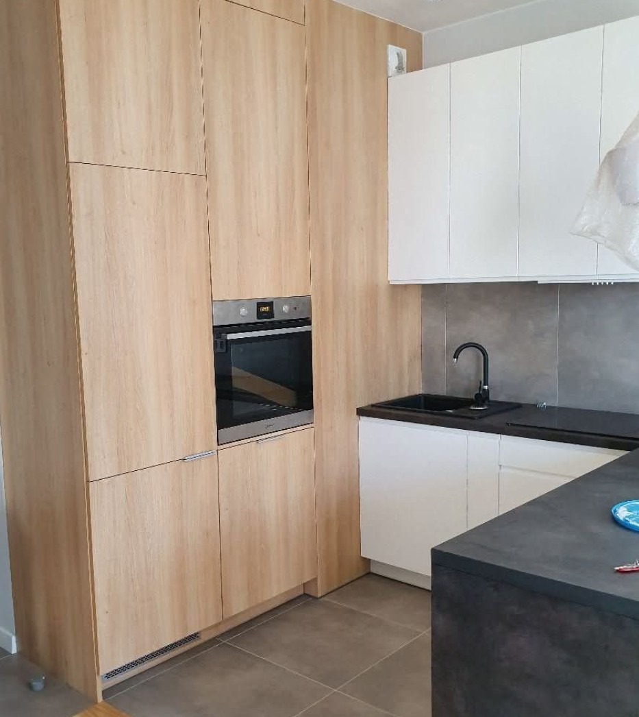 Jasna, drewniana zabudowa kuchenna z piekarnikiem w zabudowie, kontrastująca z białymi szafkami i ciemnym blatem. Nowoczesne wykończenie, szare płytki na ścianie.