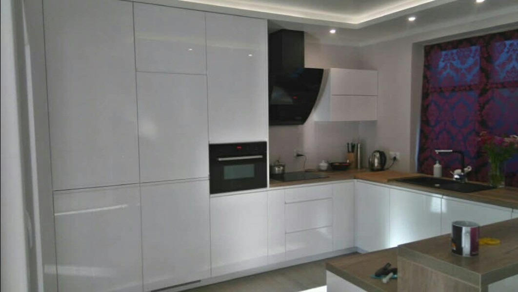 Nowoczesna, biała kuchnia z połyskiem, czarny okap i piekarnik w zabudowie. Drewniany blat i wzorzysta zasłona w tle. Oświetlenie LED podszafkowe.