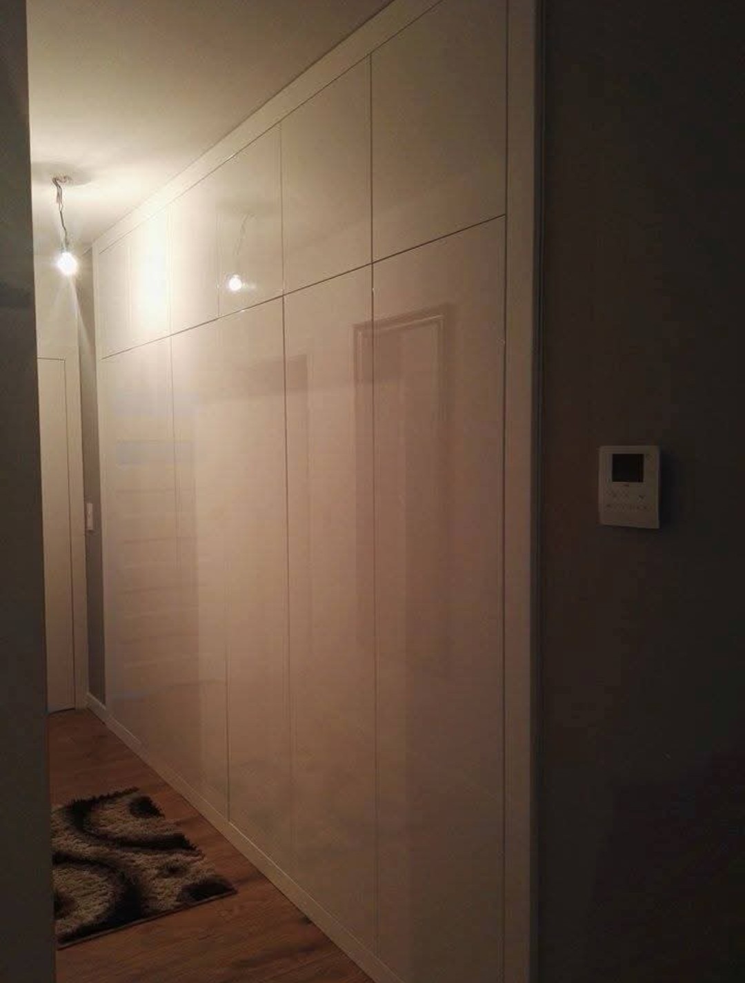 Biała szafa wnękowa z połyskiem, zabudowa od podłogi do sufitu w przedpokoju. Na podłodze dywanik, obok szarej ściany wisi regulator temperatury.