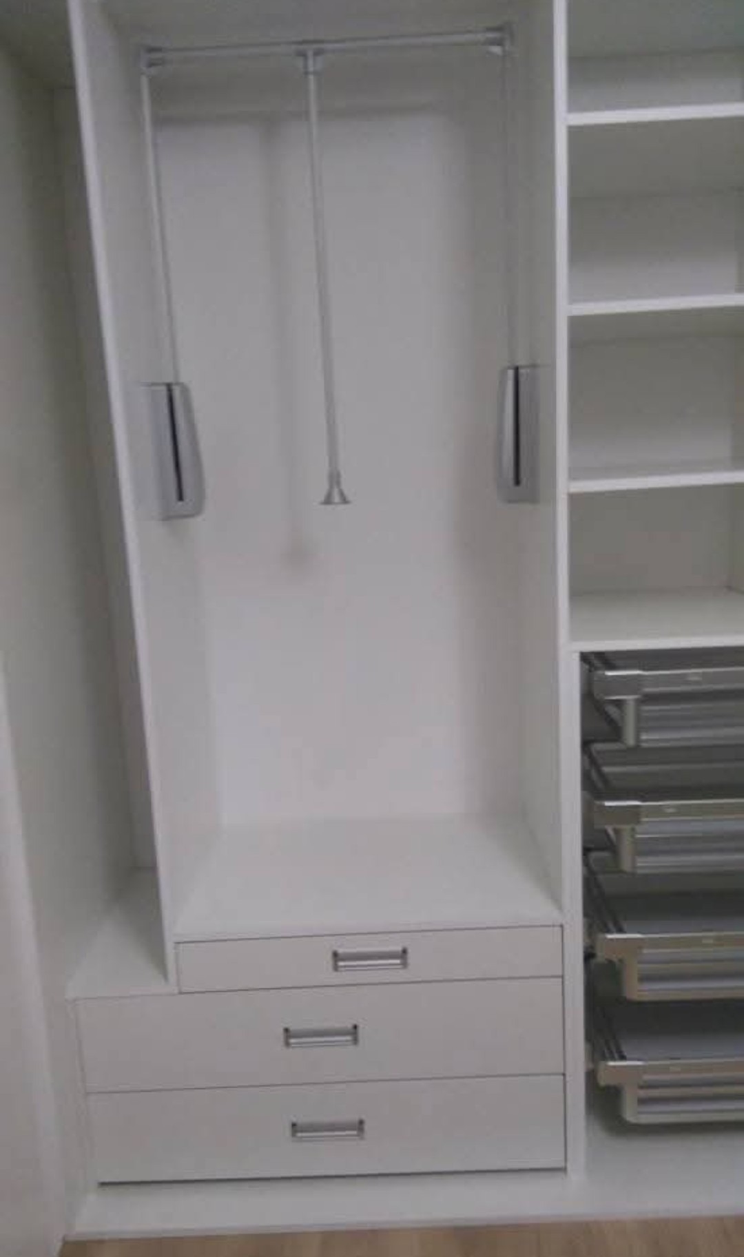 Wnętrze białej szafy wnękowej z wysuwanymi półkami i drążkiem na ubrania. Widoczne trzy szuflady z metalowymi uchwytami i półki z metalowymi koszami.