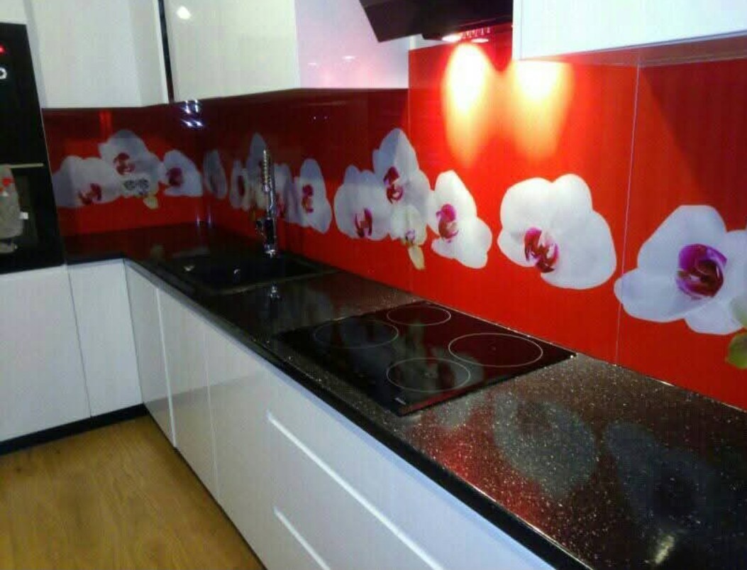Białe szafki kuchenne z czarnym, błyszczącym blatem i czerwonym panelem z motywem białych orchidei. Ujęcie na fragment zabudowy kuchennej z płytą grzewczą i zlewem.