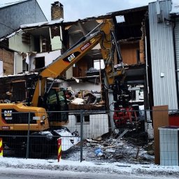 WYBURZENIA POMORSKIE MARCIN ŁATA - Żółta koparka CAT podczas rozbiórki budynku w zabudowie szeregowej, widoczne gruzy i fragmenty ścian, obok budynek kościoła.