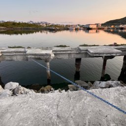 WYBURZENIA POMORSKIE MARCIN ŁATA - Betonowy pomost nad wodą z widokiem na góry i zabudowania w oddali, woda spokojna, w tle widoczne góry i zabudowania, na pierwszym planie kamienie.