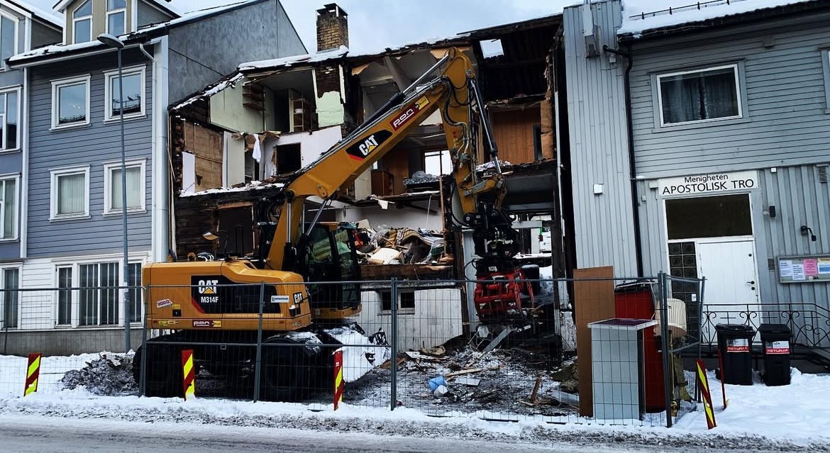 Żółta koparka CAT podczas rozbiórki budynku w zabudowie szeregowej, widoczne gruzy i fragmenty ścian, obok budynek kościoła.