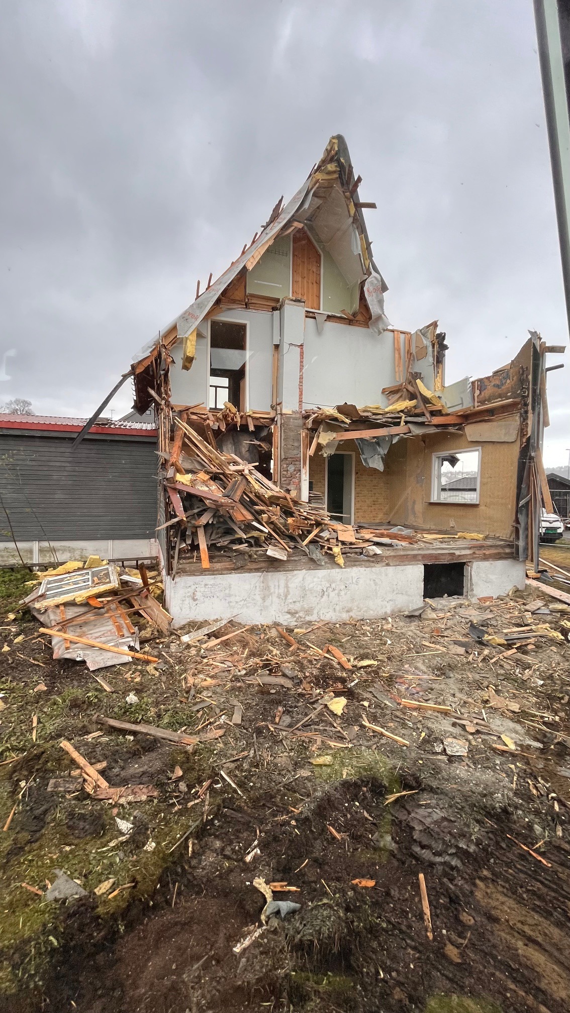 Dom po rozbiórce z częściowo zachowanymi ścianami i stertą gruzu na zewnątrz, widoczne odsłonięte elementy konstrukcyjne i zniszczony dach na tle pochmurnego nieba.