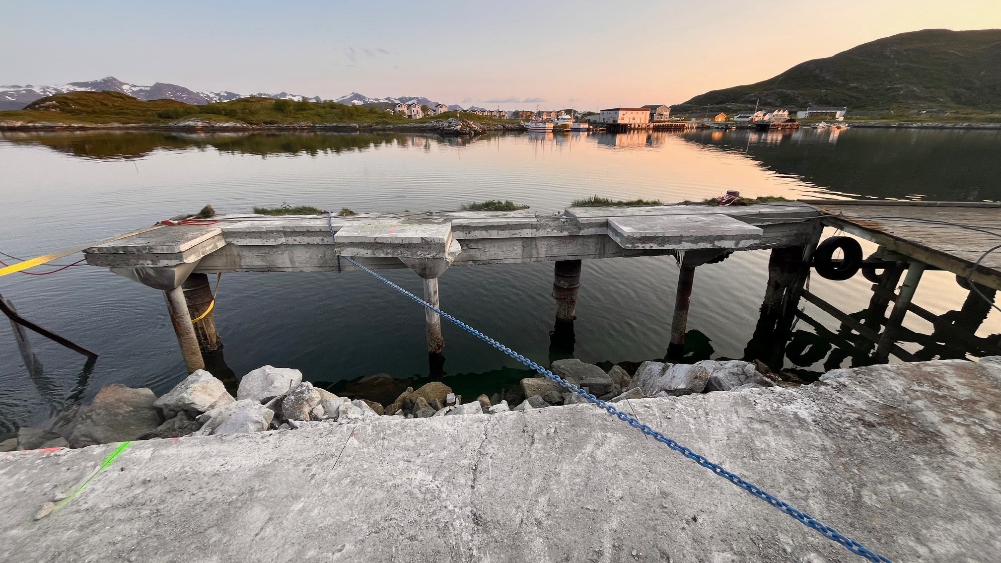 Betonowy pomost nad wodą z widokiem na góry i zabudowania w oddali, woda spokojna, w tle widoczne góry i zabudowania, na pierwszym planie kamienie.