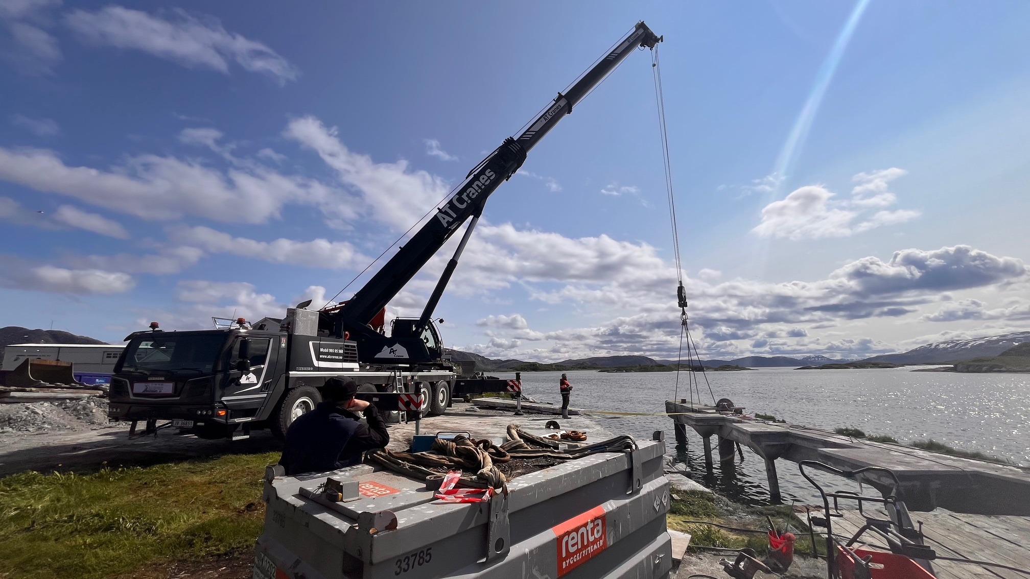 Praca dźwigu AT Cranes przy rozbiórce pomostu nad wodą. Widoczny fragment pomostu podnoszony przez dźwig, pracownicy w oddali i krajobraz z górami w tle.