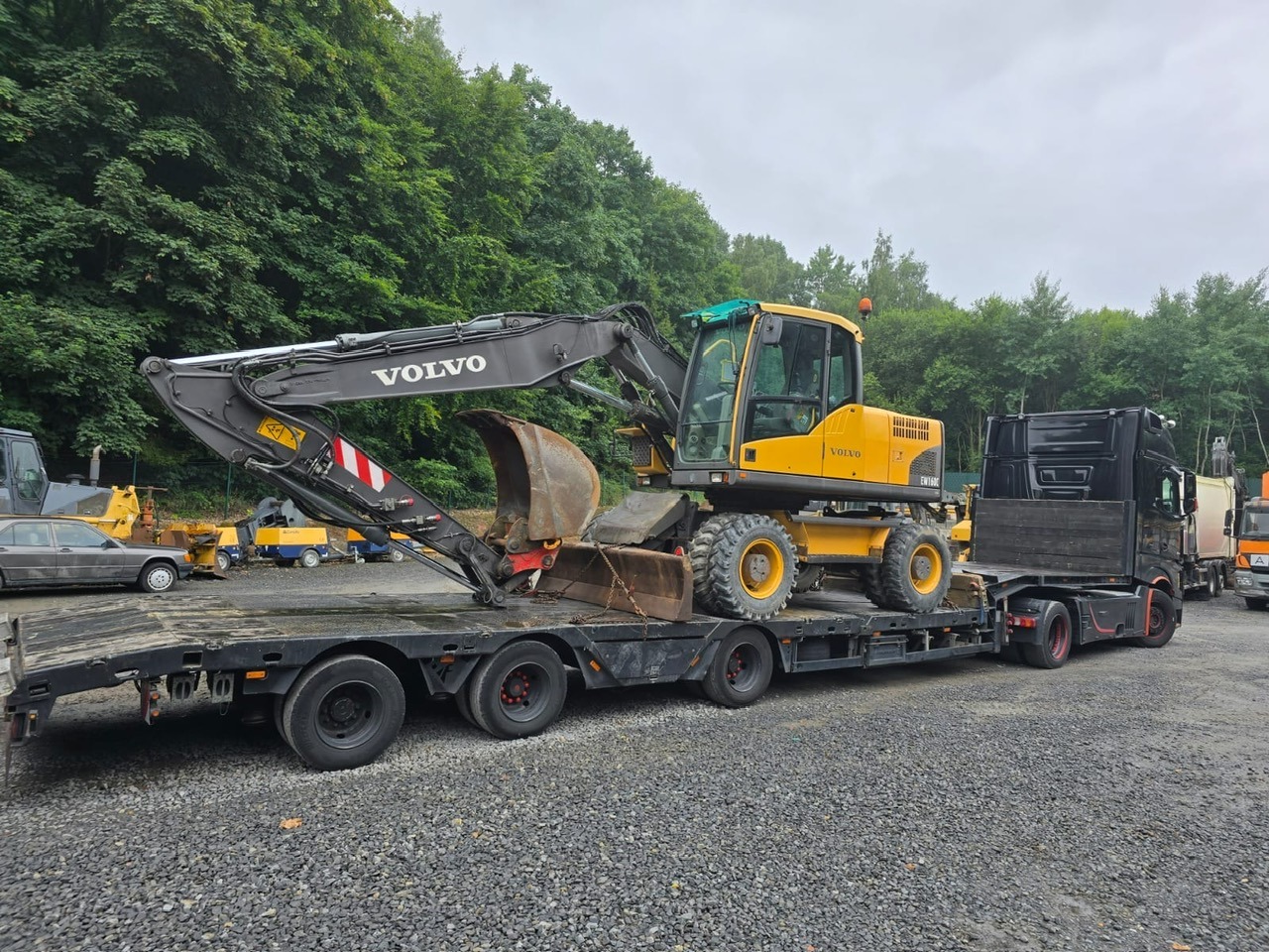 Żółta koparka Volvo EW160C na czarnej lawecie, transportowana przez ciężarówkę. W tle drzewa i inne maszyny budowlane na żwirowym podłożu.