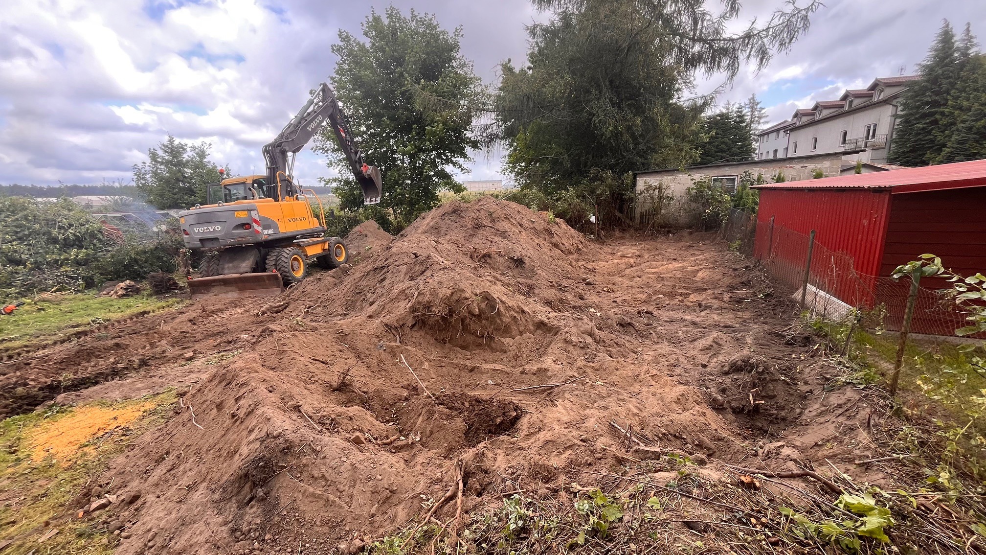 Żółta koparka Volvo na terenie budowy, hałda ziemi w wykopie, widoczny budynek w tle. Prace ziemne w toku, przygotowanie terenu pod budowę.