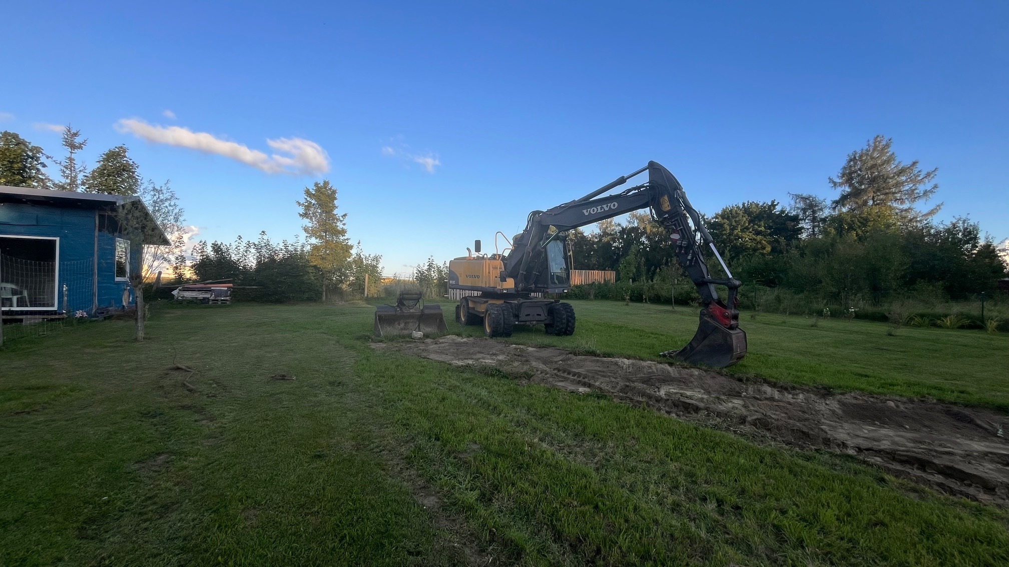 Żółta koparka Volvo na trawniku, w tle domek z niebieską elewacją. Wykopany wąski pas ziemi. Dwa łyżki obok maszyny. Błękitne niebo.
