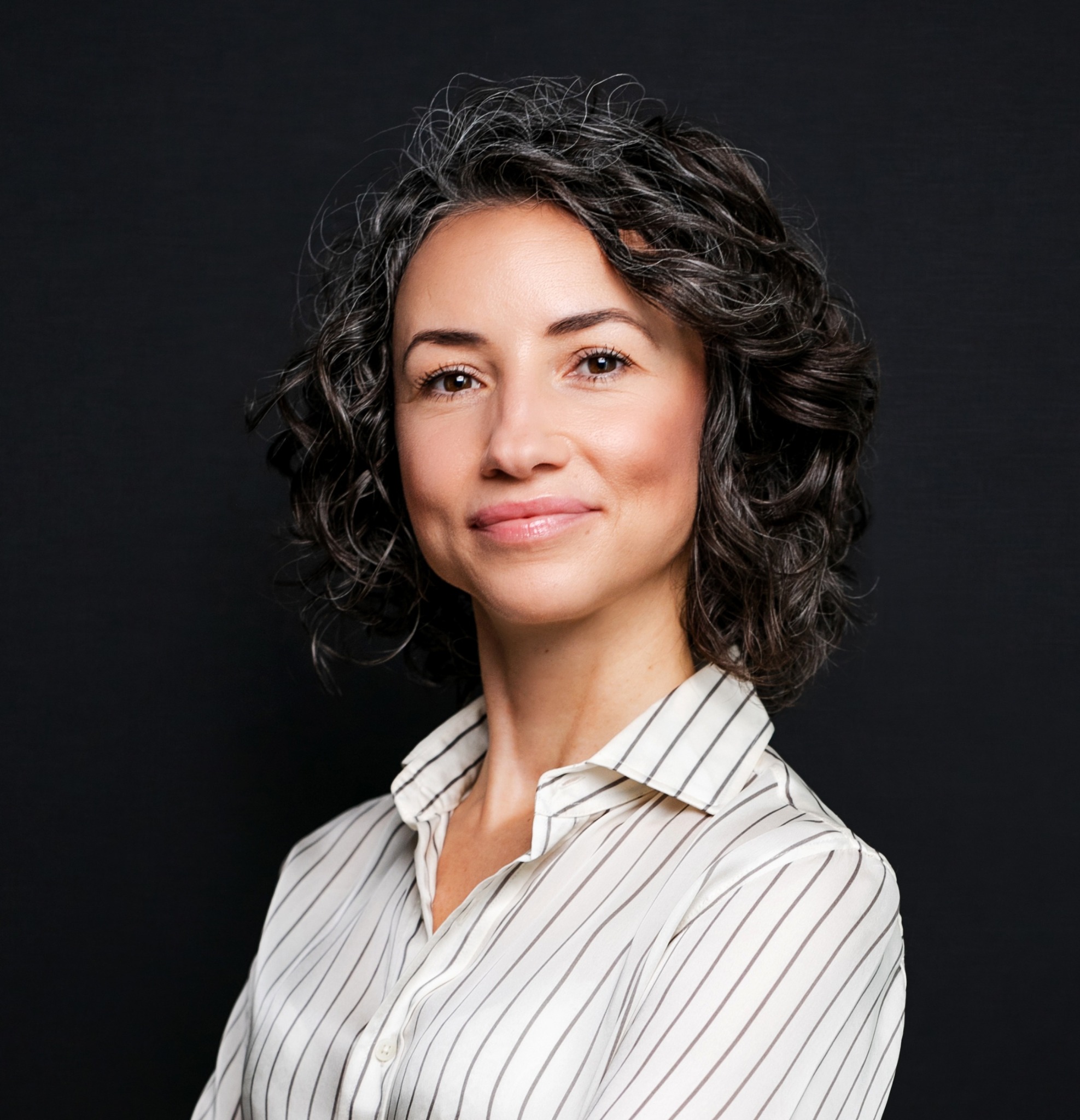 Portret kobiety o kręconych, siwiejących włosach w bluzce w paski na ciemnym tle. Uśmiech, naturalny makijaż, profesjonalny wygląd.