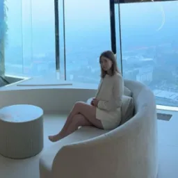 Profesjonalistka w białym garniturze siedzi na sofie w nowoczesnym wnętrzu z widokiem na miasto. Jasne kolory i minimalistyczny design biura.