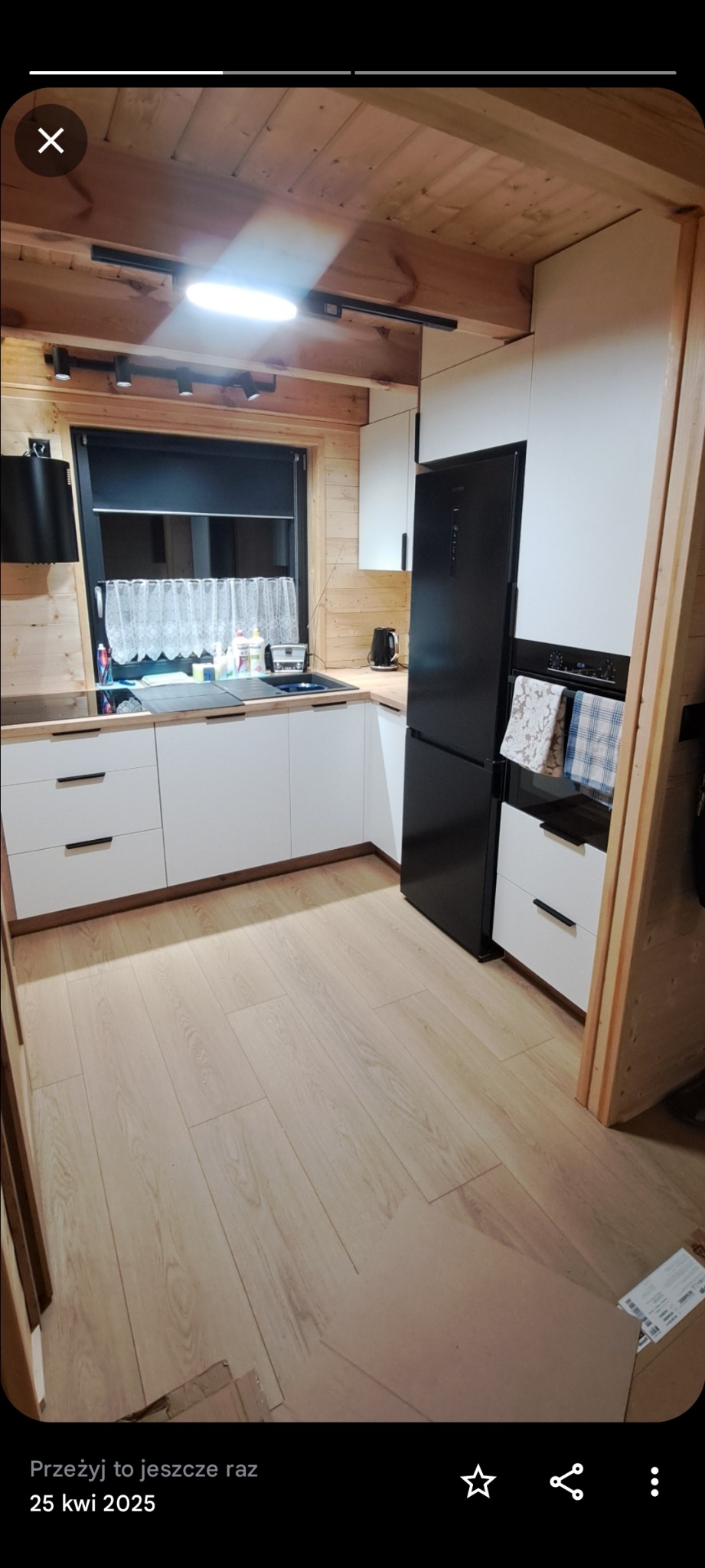 Jasna kuchnia w stylu skandynawskim z drewnianym wykończeniem, czarnym sprzętem AGD i białymi frontami szafek. Widoczne oświetlenie szynowe i drewniana boazeria.