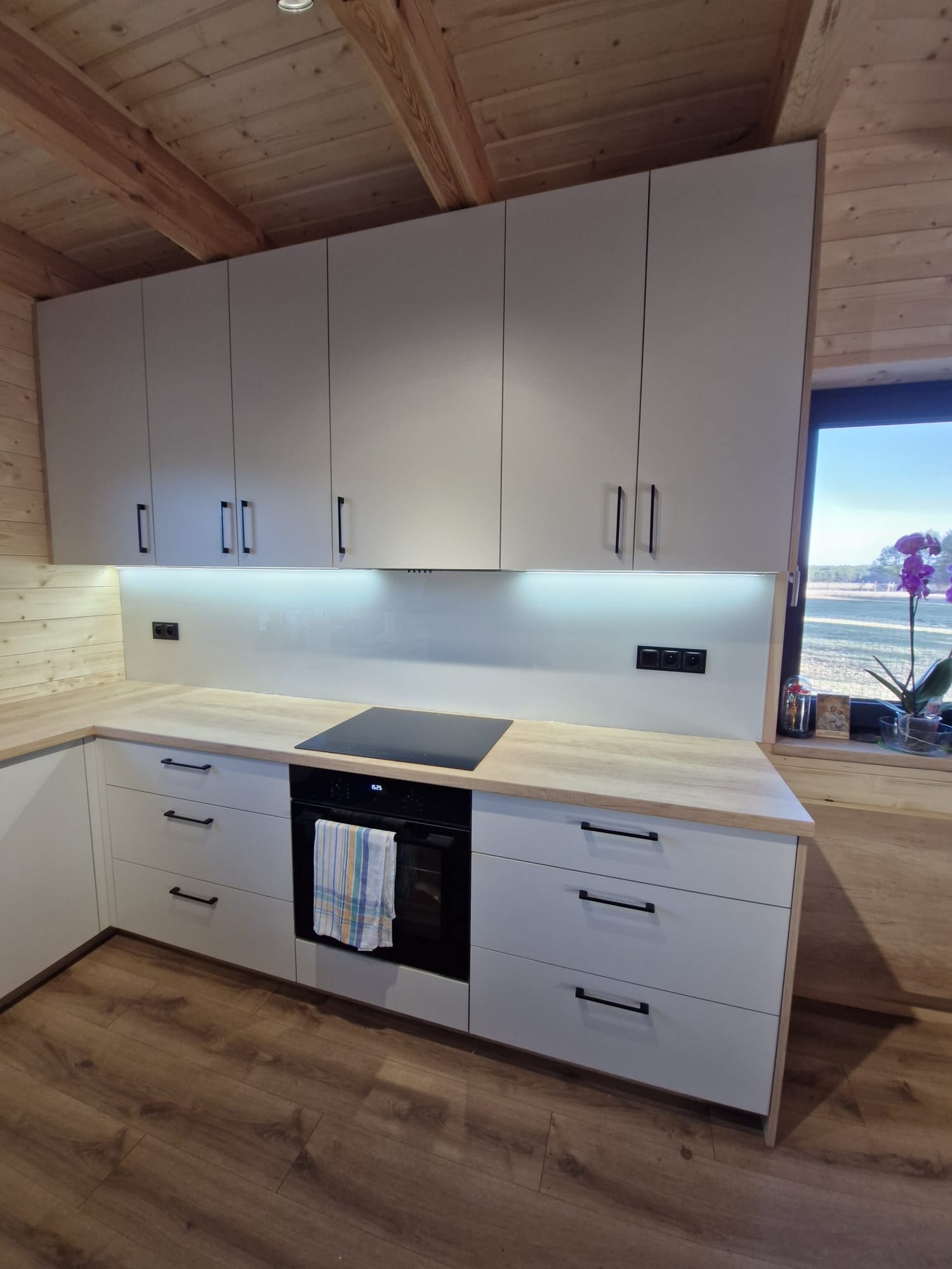 Jasna kuchnia w domku z drewnianym stropem. Białe szafki z czarnymi uchwytami, drewniany blat, płyta indukcyjna, okno z widokiem na zewnątrz i kwiaty.